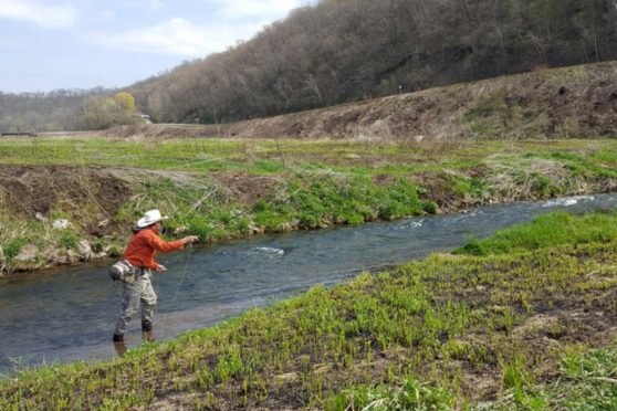 An angler fishing Prairie Song Farm in northeast Iowa. Note the sloped banks and recently-burned prairie grass starting to regrow after the annual spring burning.