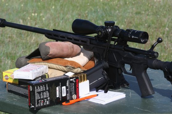 Black finish Howa chassis rifle with open bolt and scope, resting on shooting bags with open box of Winchester .308 ammunition nearby and shooters log notepad.