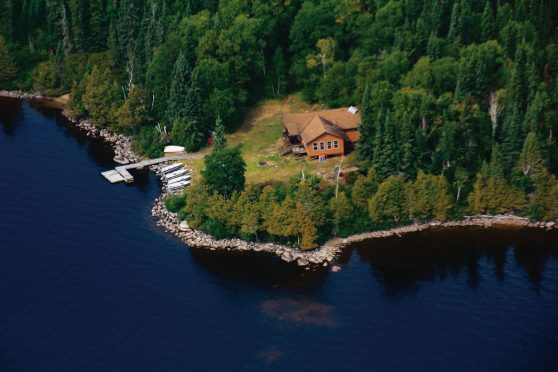 Aerial photo of a brown-roofed fishing cabin, Wabakimi Lake Outpost, on a heavily wooded point next to a rocky breakwater point.