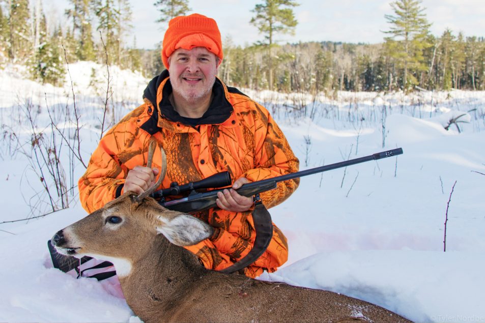 Deer hunter in the snow with small "spike buck" deer and rifle.